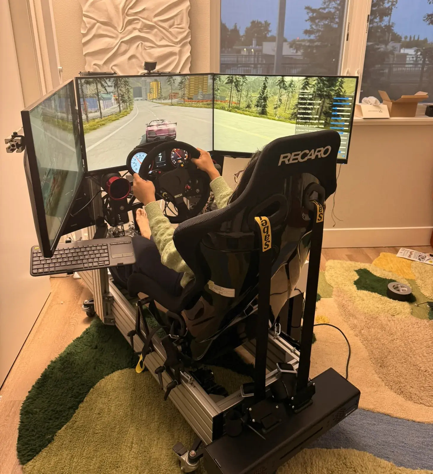 My racing sim at dusk: the Recaro seat on its aluminium rig, the wheel, and a road across its three screens.